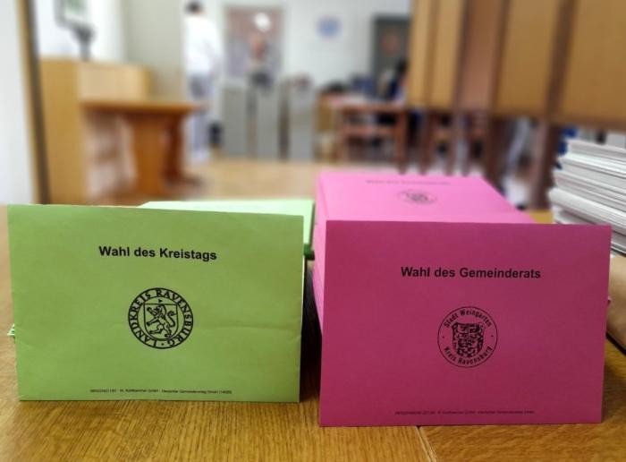 Ein Stapel Briefumschläge für die Wahlen in den Farben Rot und Grün, im Hintergrund Blick ins Wahllokal im Haus am Mühlbach 
