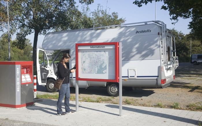 Wohnmobilstellplatz Eine Frau mit Handy steht am Stadtplan der Infotafel, im Hintergrund ihr Wohnmobil auf dem Stellplatzes