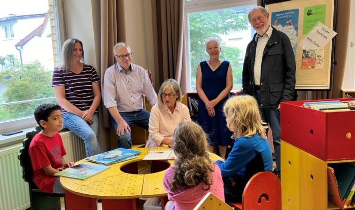 Kinder sitzen an einem Tisch, eine Frau liest aus einem Buch vor, im Hintergrund Vertreter der Bürgerstiftung und der Bücherei Weingarten.