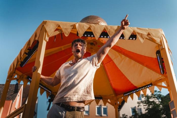 Auf einer Bühne mitten in der Stadt schreit ein Schauspieler mit ausgestrecktem Arm und weit aufgerissenem Mund