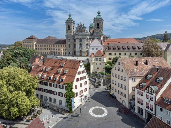 Häuser, ein gepflasterter Platz mit rundem, weißem Bodenmosaik sowie die Basilika hoch über der Stadt
